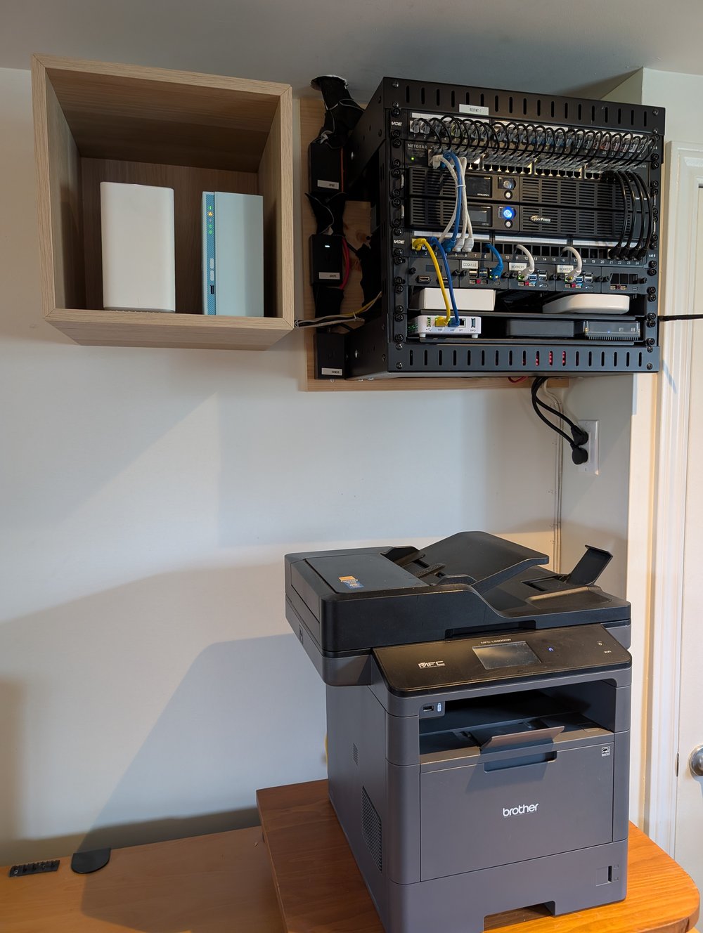 Coin du sous-sol : le rack mural avec les trois Raspberry Pi, un cube mural IKEA à gauche contenant le NAS et un nœud Helix Fi, et l’imprimante multifonction Brother sur une table en dessous
